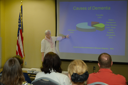 Presenter during the 2013 seminar. Audience is looking at presenter who is pointing to a PowerPoint slide titled Cause of Dementia that has a pie chart with the different causes and percentages.