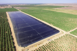 Arial image of an empty oblong reservoir lined with a dark material. The reservoir is surrounded by fields of trees and tiny trucks can been seen in the foreground, revealing the enormous size of the reservoir.