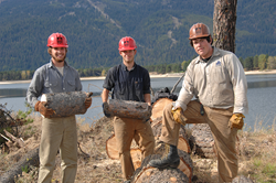 Three young men stand with logs in front of Lake Cascade Reservoir.