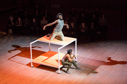 Surrounded by audience, two dancers on a 2 level platform on wheels.