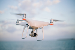 Blue, cloudy sky and water background blurred, with white drone midflight in focused foreground, silver propelling blades spinning, camera attached.