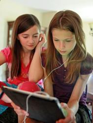 Two young girls with dyslexia share earbuds while listening to a Learning Ally audiobook on the iPad
