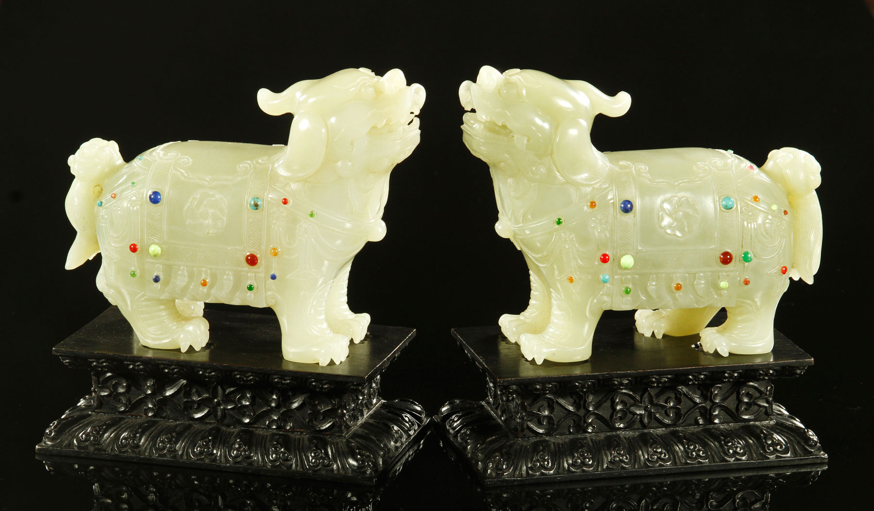 Carved pair of jade foo lions, China, in standing pose and carved wearing garments inlaid with colored semi-precious stones, on hardwood stands, 5" x 6" x 2 1/2", on stand, 7" h.