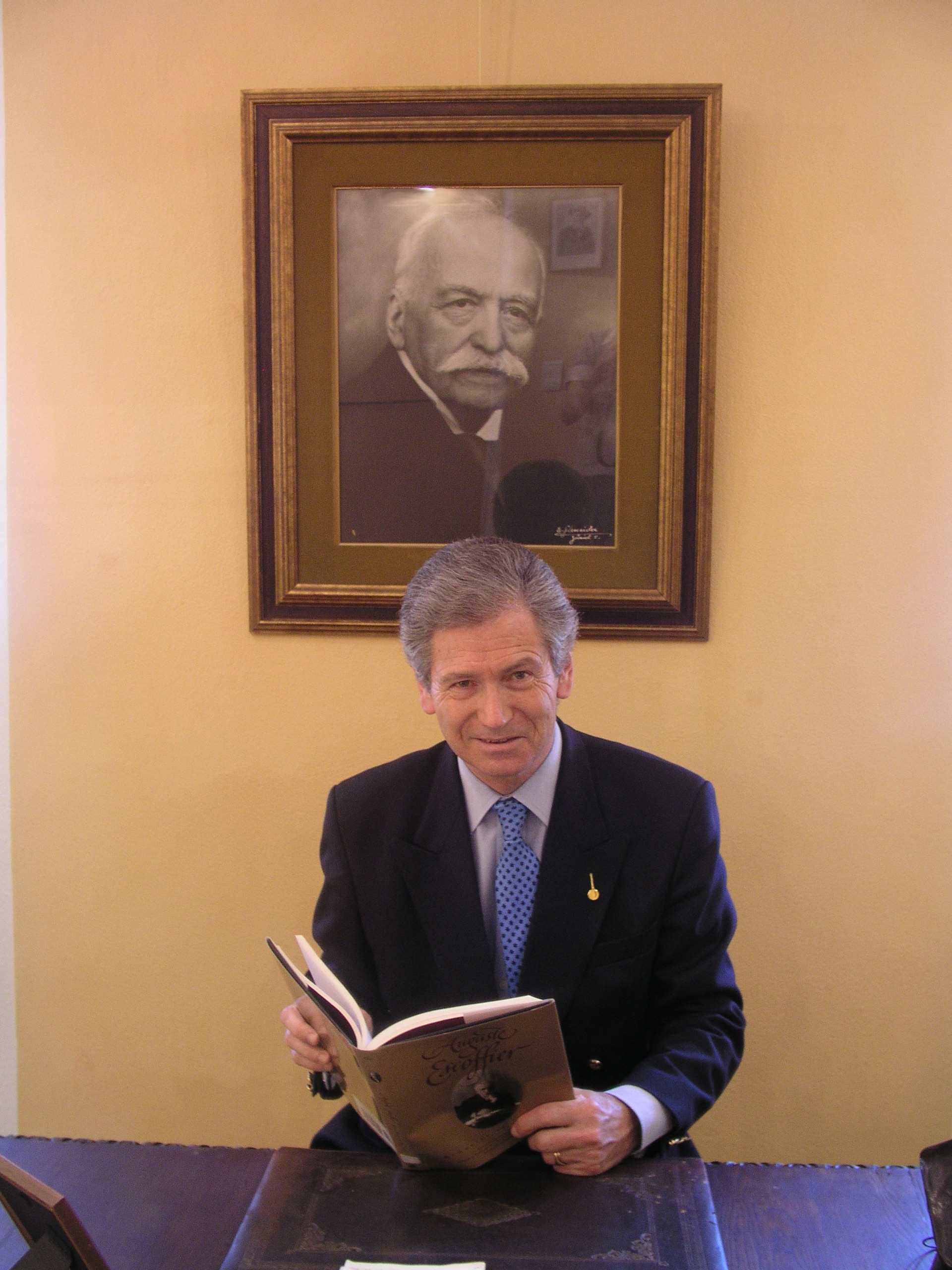 Michel Escoffier seated in front of a photo of his Great Grandfather Master Chef Auguste Escoffier