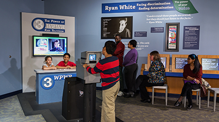 Children and families pretend they're newscasters talking about AIDS/HIV and Ryan White in The Power of Children exhibit