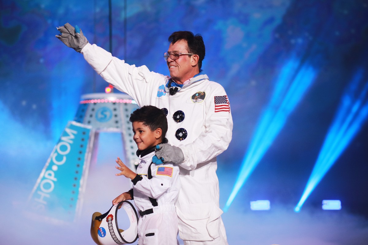 Ayden McLaughlin & JR Ridinger, dressed in spacesuits, emerge from a NASA space capsule that landed onstage to visually demonstrate that The Shopping Annuity landing on Earth.