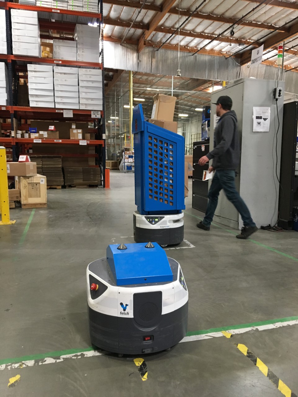 An RK Logistics associate walks by as two Fetch Robotics autonomous delivery robots arrive. The robots move completed orders from assembly to shipping, relieving employees of tedious tasks