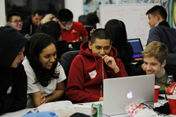 Four high school students huddle around a laptop, looking happy.