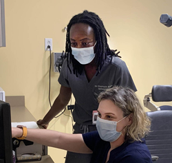 Dr Stephanie Aigbe is standing and looking over Dr. Lexi Malkin's shoulder at a patient record in preparation of a visit.
