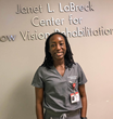 Dr. Stephanie Aigbe is standing below a sign for the Janet LaBreck Low Vision Rehabilitation Center at the New England College of Optometry.