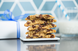 A stack of chocolate chip cookies sits on a small plate in front of a glass of milk and a Tiff's Treats-branded white delivery box tied with a blue ribbon.