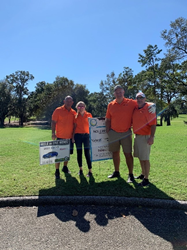 Three men and one woman stand together on a golf course with trees in the background. The sky is bright blue and cloudless, and the day is bright and sunny. They're standing next to a yard sign showing event sponsors and all wearing orange polo shirts.