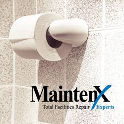 A roll of toilet paper on a white toilet paper holder, against a grey and white specked tile wall. At the bottom right of the image is the MaintenX International logo.