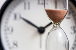 An hourglass with sand inside in front of a clock.