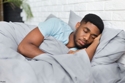 Man in light blue t-shirt sleeping in bed with gray bedsheets.