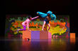 Two actors on stage atop pink, purple and orange square blocks. One actor kneels with her arm outstretched toward the other actor who is wearing a blue, scruffy monster costume. Her arms are outstretched to scare the other actor.