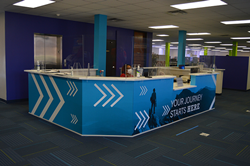 A welcome desk with decals depicting a mountain and mountain climber. There are green arrow graphics placed throughout. The background is a blue sky.