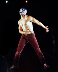Irvin Manuel Gonzalez is captured in a moment of performance. He stands before a black background, looking up to the left corner. Irvin wears a luchador mask, no shirt, and maroon pants.