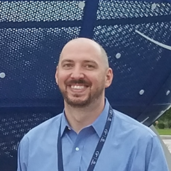 A smiling bald white main is standing outside. He has dark eyes and dark facial hair and he is wearing a light blue button-down shirt.