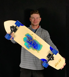 Keaton Kissak stands in front of a black background, arms outstretched, holding a longboard with bue wheels. It is made of raw wood, with a laser-cut topo map of Lake Tahoe painted in variegated blue.