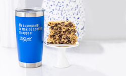 A blue YETI Rambler tumbler reads, "My superpower is making cookies disappear" with a stack of freshly baked chocolate chip cookies sitting on a small cake stand in the background.