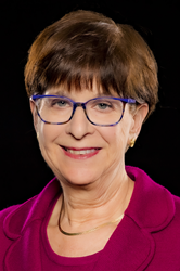 White woman with short brown hair and blue glasses, wearing gold necklace, and bright pink round collar sweater and matching jacket.