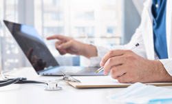 A physician consults notes from a clipboard while updating information on their laptop.