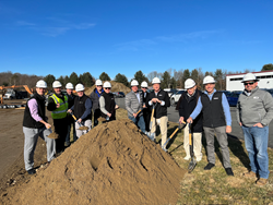 A group of smiling people wearing PPE hold shovels around a large pile of dirt.