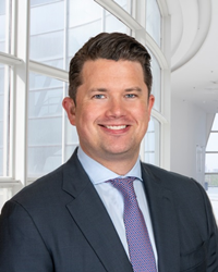 A white man wearing a gray suit, blue shirt and tie with short brown hair is smiling while standing in front of a bank of large windows.