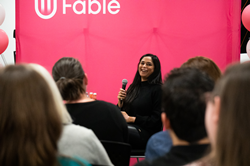 Photo of Alwar sitting in front of a Fable backdrop, holding a microphone. Foreground of team sitting watching her. Alwar smiles.