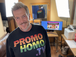 Nicholas Snow stands close to the camera with his professional home broadcast studio in the background. Snow is wearing an Official PromoHomo.TV logo sweatshirt in black. In the background are two brightly lit studio lights and an l-shaped desk with a wide screen computer monitor and printer visible. Snow has a short salt and pepper beard and is smiling.