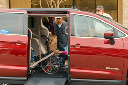 A young girl in a wheelchair is secured in a red wheelchair van with a side-entry ramp made by BraunAbility. Her family was granted the vehicle as part of a partnership between Laughing at My Nightmare and BraunAbility.