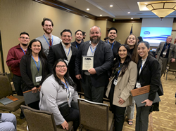 University of La Verne Assistant Professor Russell Muir holds award standing with students and colleagues in a conference room.