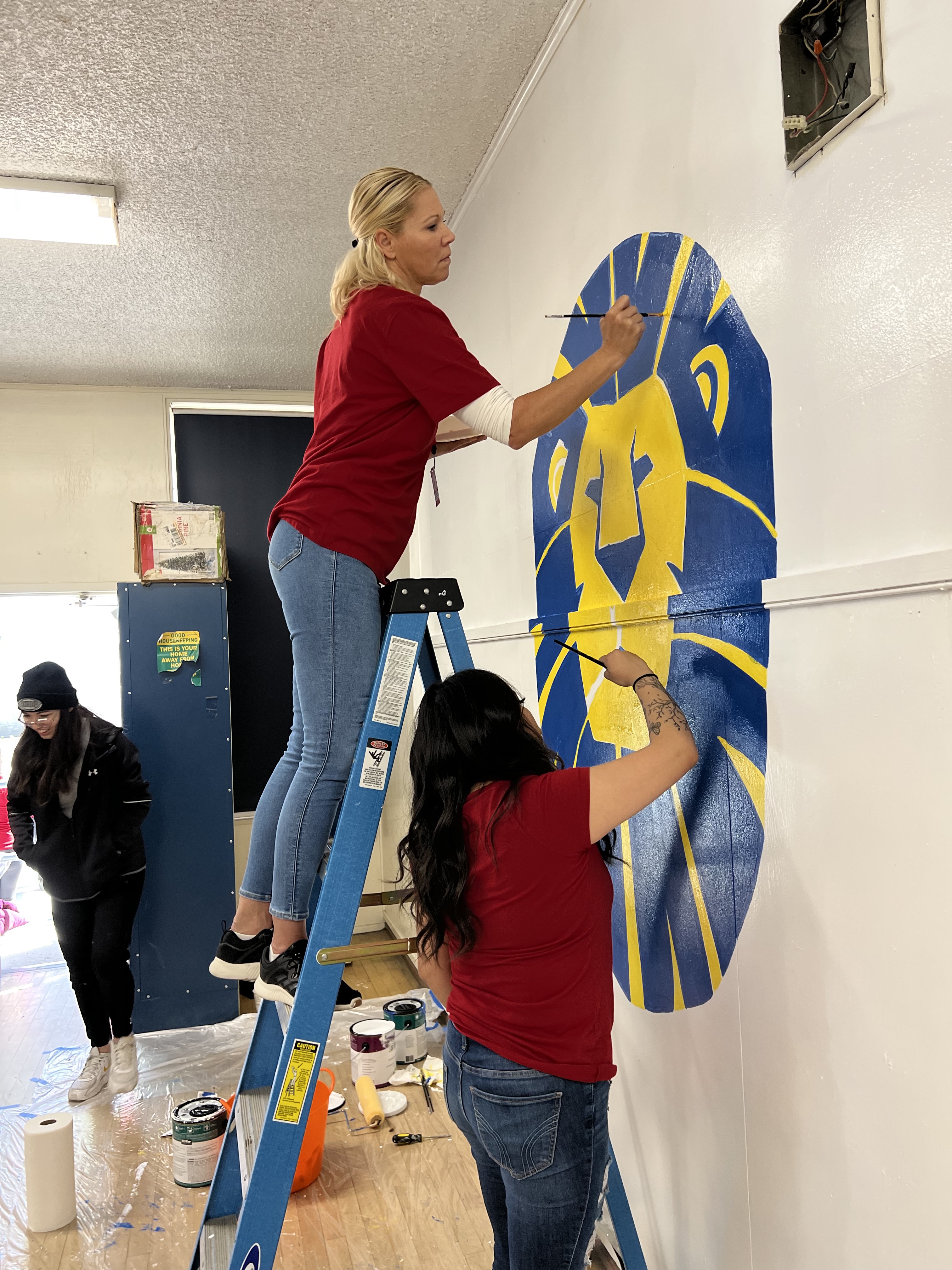 Volunteers paint a mural of the mascot on the cafeteria stage wall.